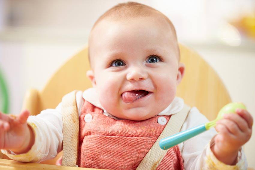 A happy baby sitting on a chair with a spoon in their hand