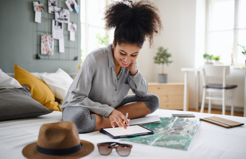 Person preparing for a trip, using a tablet with a map underneath it and travel documents spread out on a bed in a bright bedroom.
